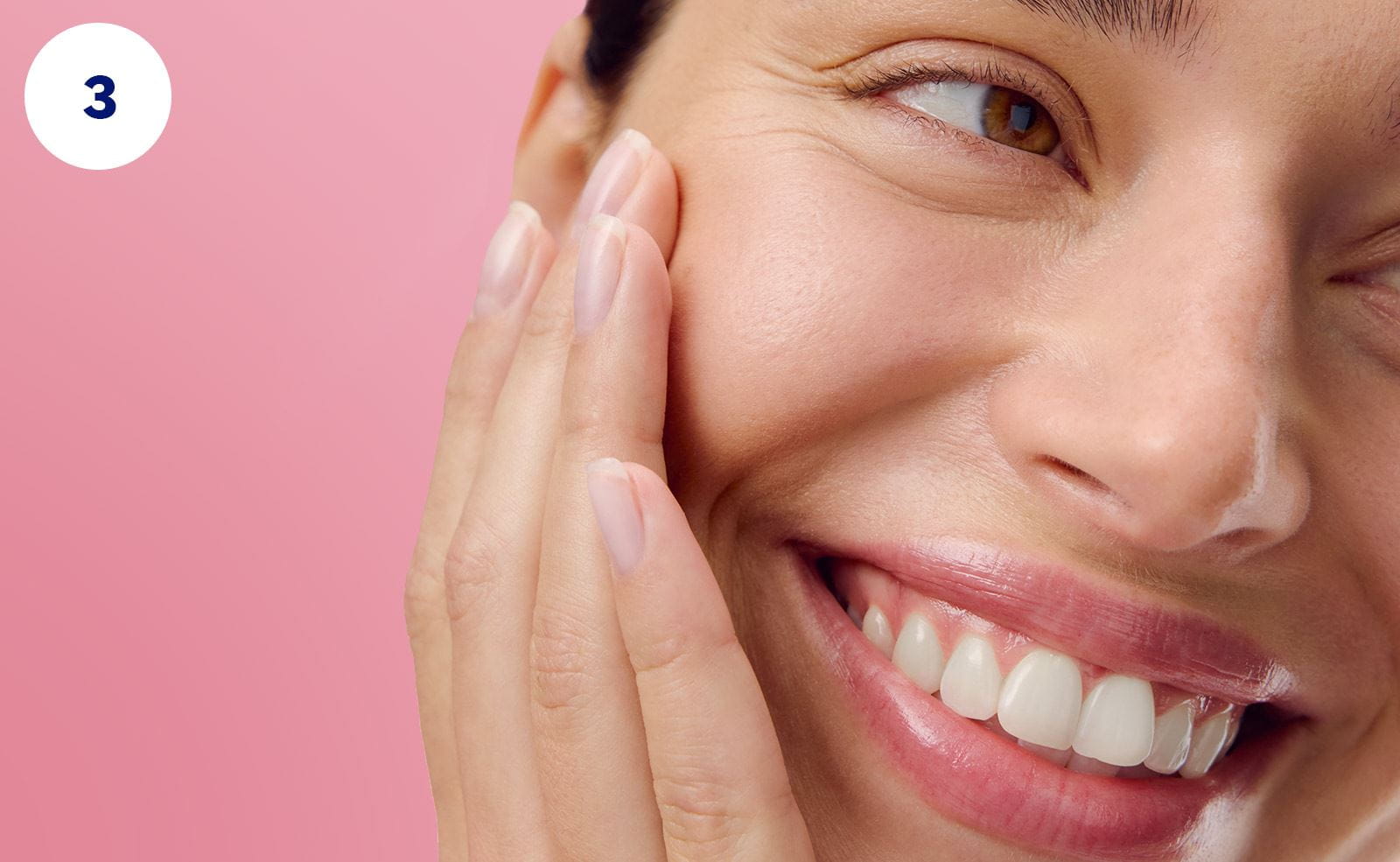 A close-up of a person smiling while holding their face in their hand, against a pink background.