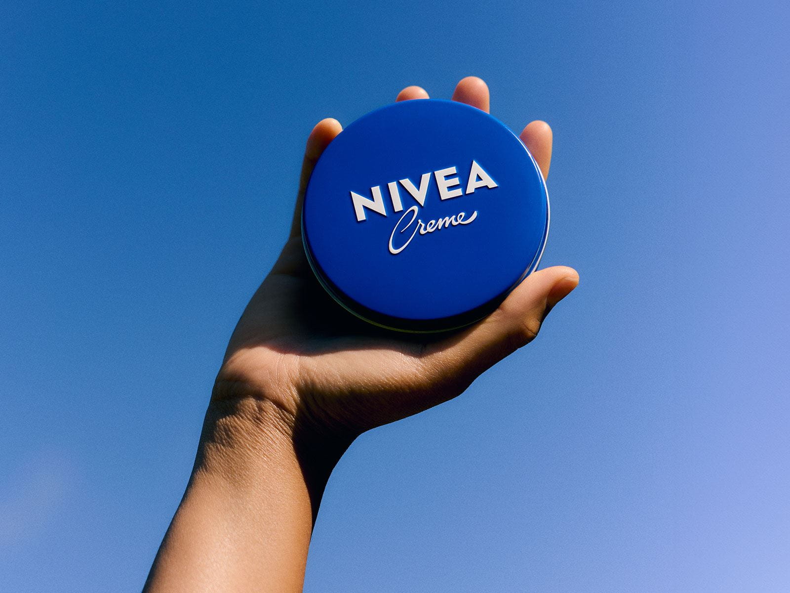 Le produit NIVEA Creme bleue tenue en l'air, sur fond de ciel bleu.