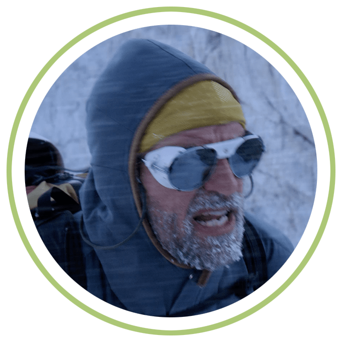Image of a man hiking during snowfall, his beard is covered in frost. He’s dressed in heavy winter gear with protective eyewear.