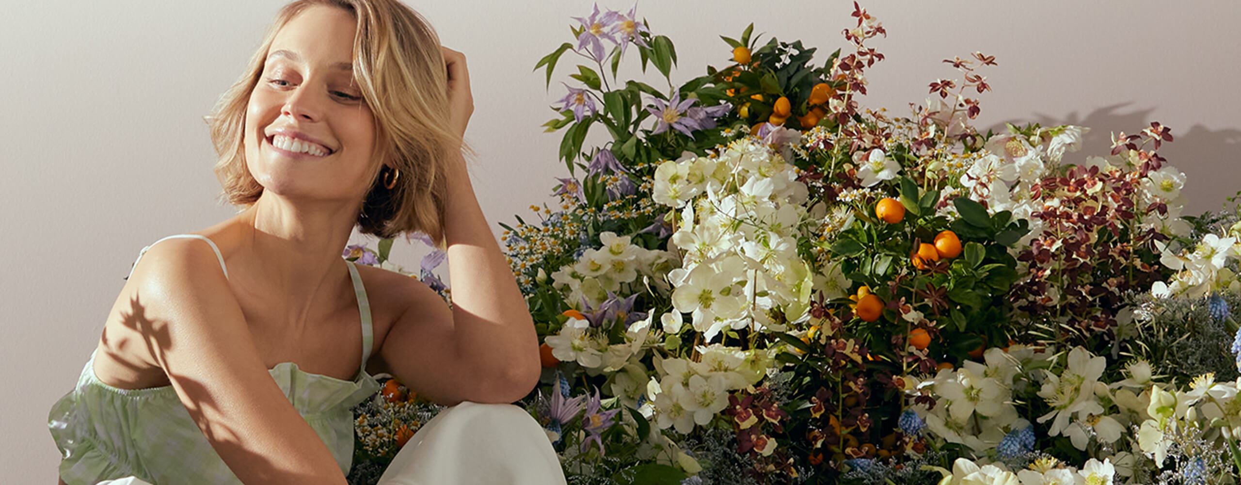Vue d’une mannequin aux cheveux blonds courts regardant vers le bas, avec un arrangement floral derrière elle.
