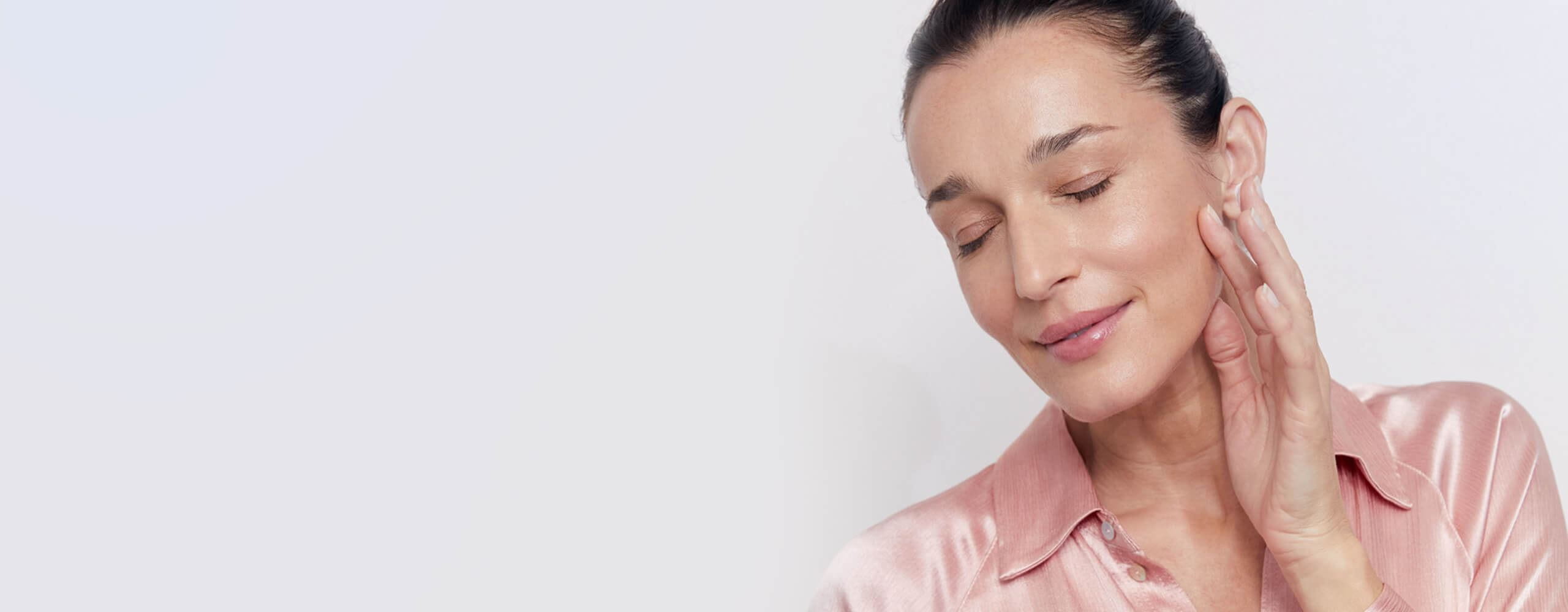 View of a female model with brown hair and wearing a light pink silk shirt closing her eyes against a light grey background.