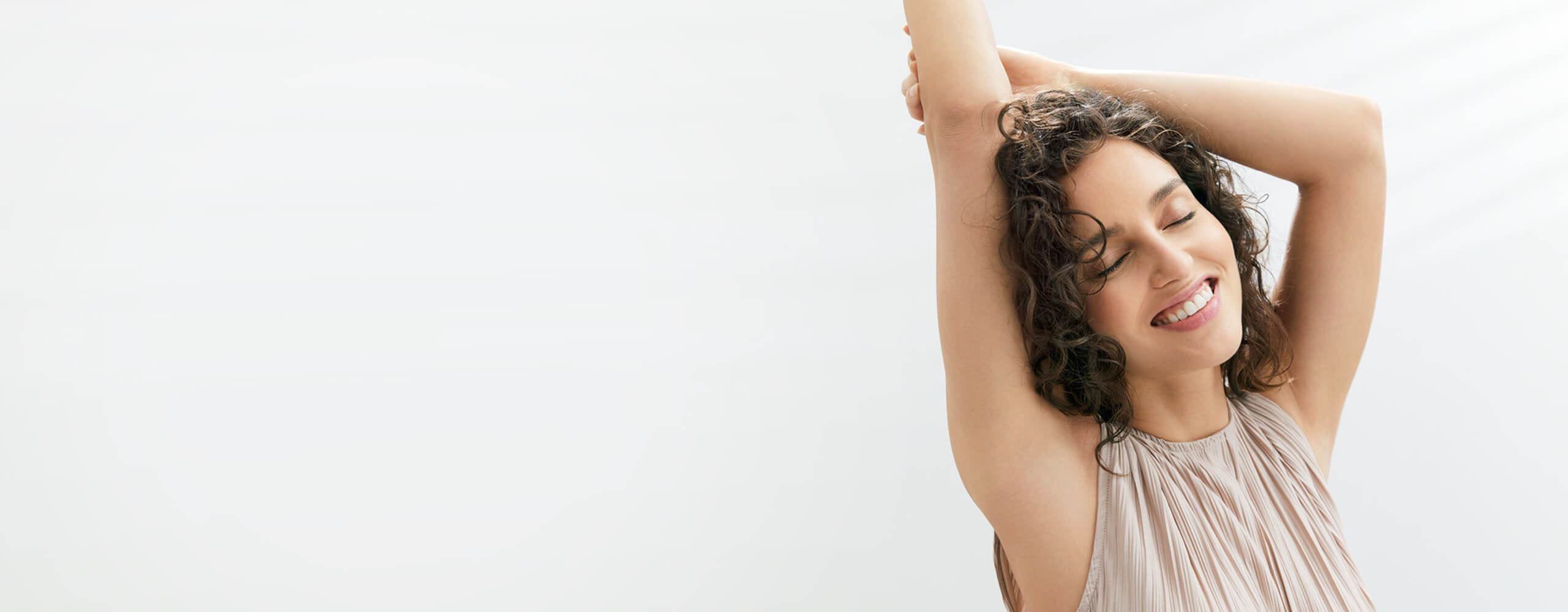 View of a female model with brown curly hair closing her eyes and raising her arms above her head against a grey background.