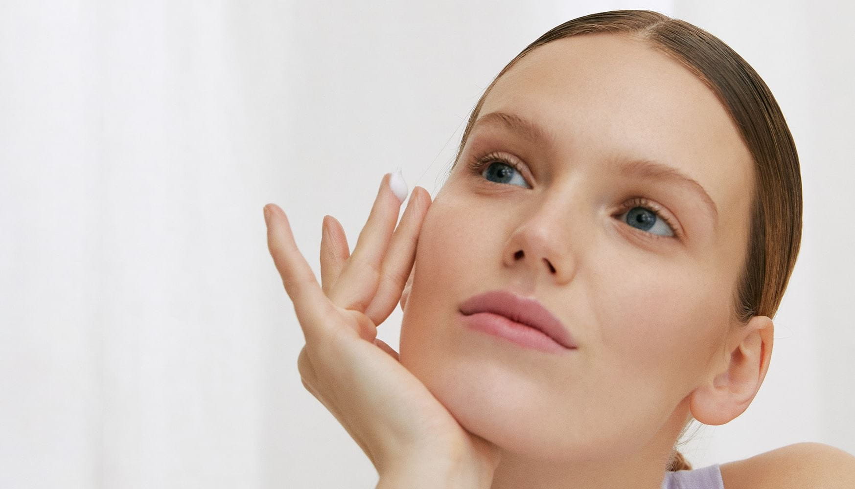View of model with right hand pressed on the face and product smeared on ring finger against a white background.