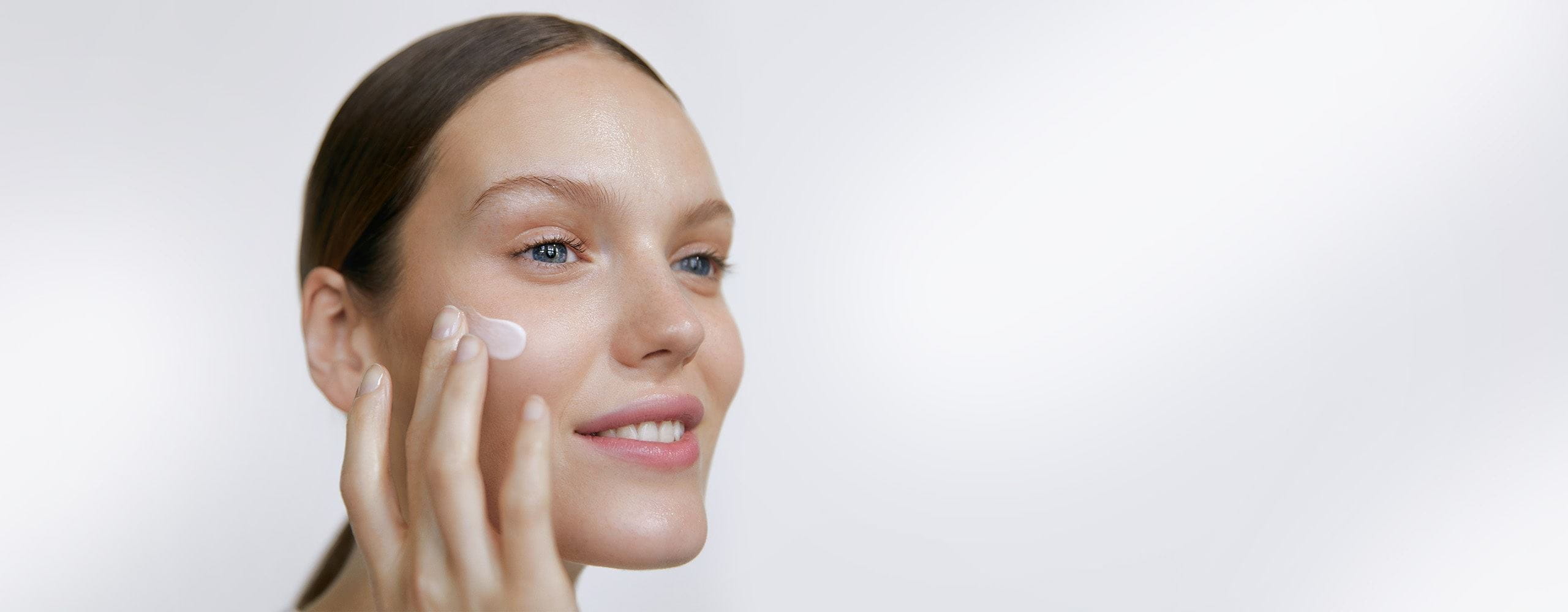 Vue d'un mannequin souriant appliquant un produit avec la main droite sur le côté droit du visage sur un fond blanc.