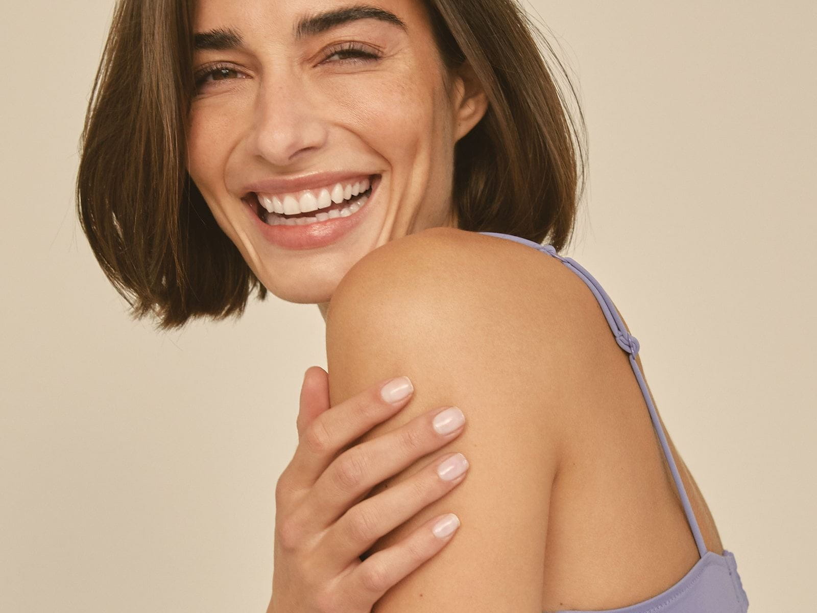 View of a person wearing a light coloured tank top while smiling and touching their left shoulder.