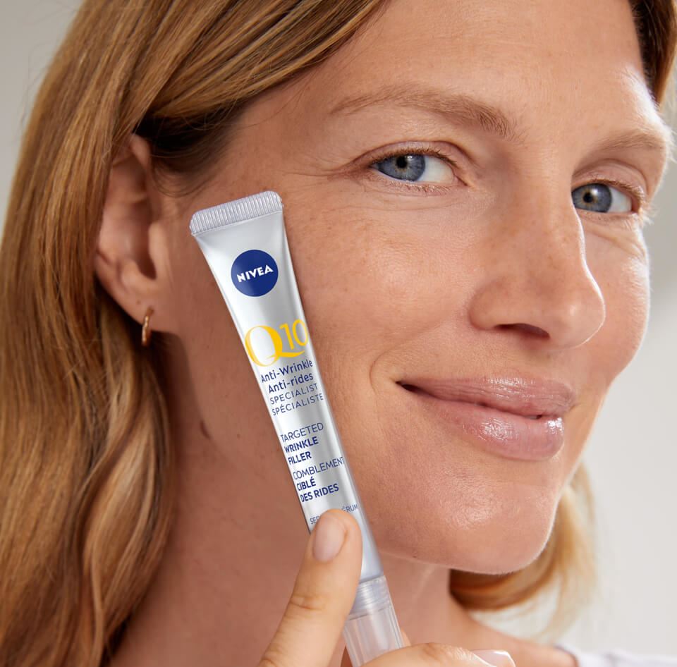 View of a female model smiling while holding up the Nivea Q10 Anti Wrinkle Specialist targeted wrinkle filler product against her cheek.