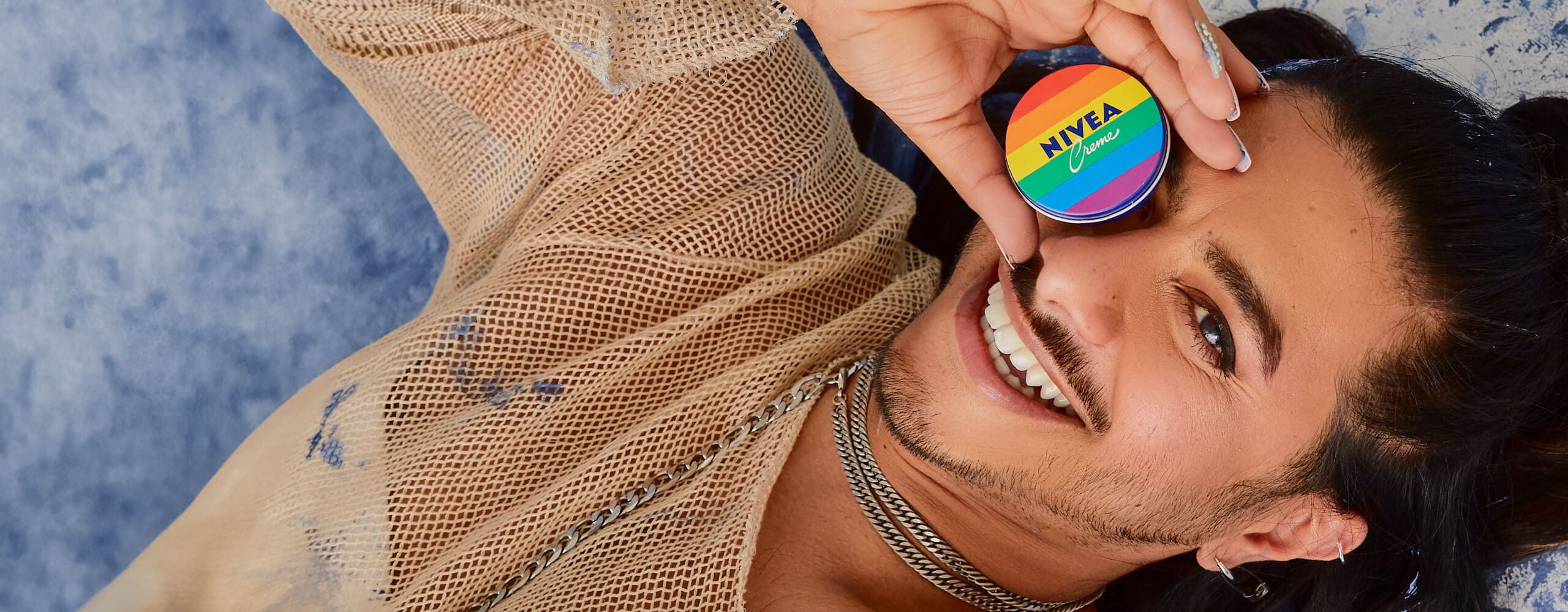 A view of a person laying down and smiling, while holding a rainbow-coloured Nivea Crème Pride edition product against their face.