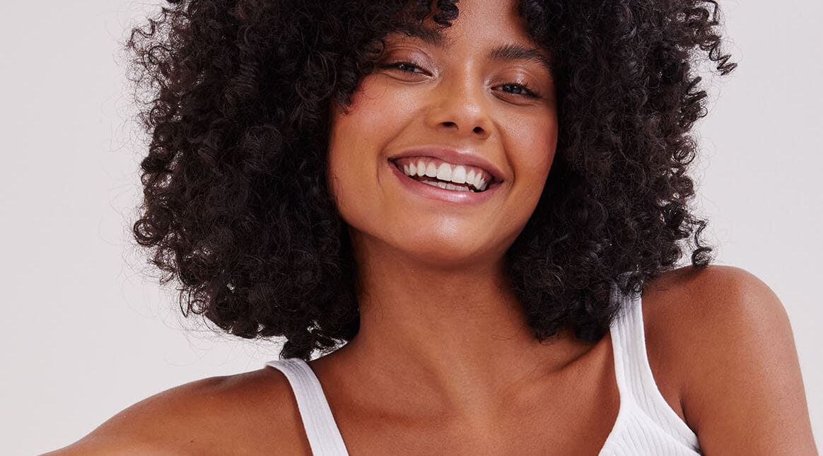 A view of a person with dark-coloured short, curly hair smiling against a white-coloured background.